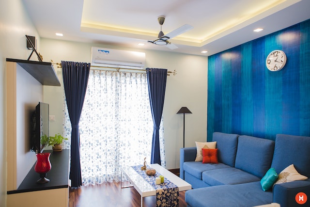 a living room with a blue accent wall  with a blue sectional and a square white coffee table facing a television natural light streams in through the white curtains and lights up the space